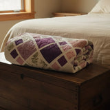 Folded handmade modern heirloom king quilt in shades of lavender and green on a wooden chest at the foot of a bed