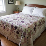 Lavender and green handmade quilt on a wooden bed with warm farmhouse decor, brass lamps, and gentle window light.