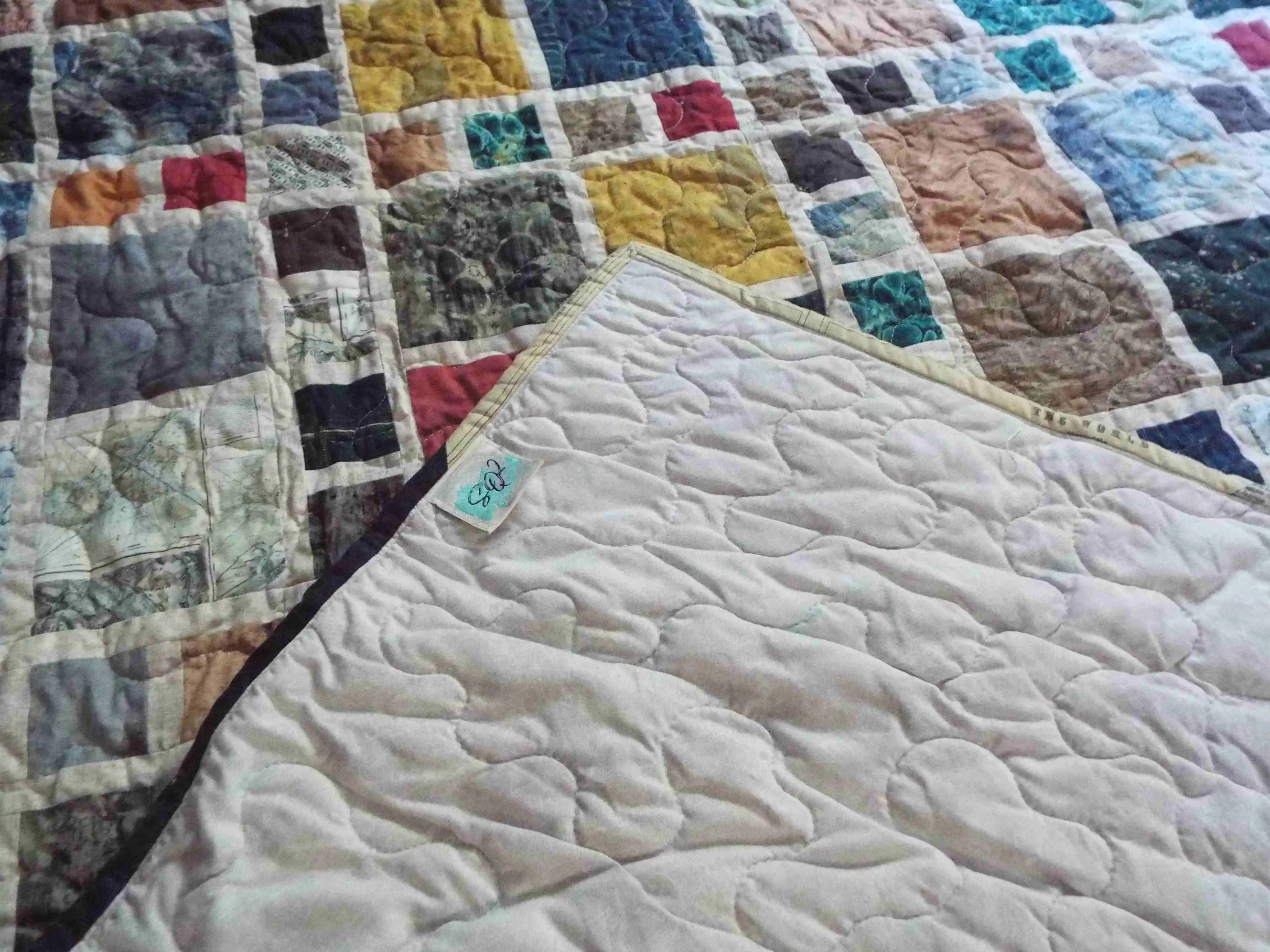 Corner of the Of Ink and Thread King Quilt folded to reveal the soft neutral backing, signature SQ² label, and free-motion quilting pattern.