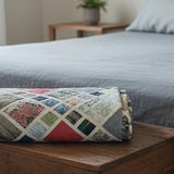 Folded handmade king quilt in layered blue, rust, and parchment tones resting on a gray wooden chest at the foot of a bed.