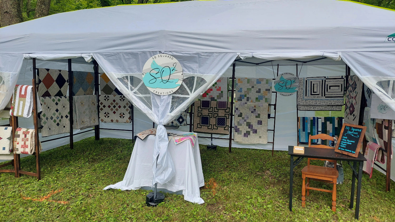 Quilt show booth with various quilts on display under a white canopy.