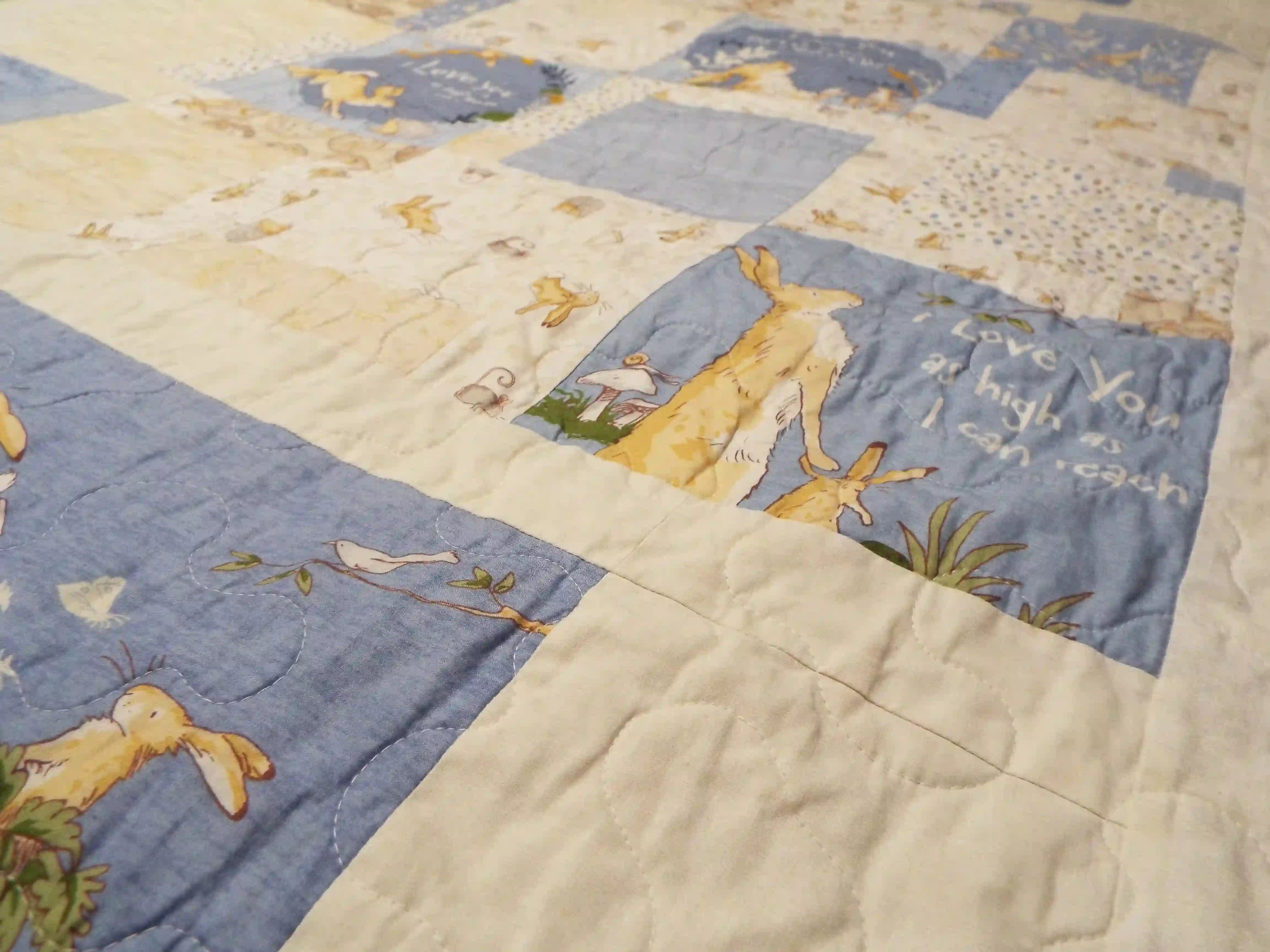 Angled close-up of a handmade quilt in blue and tan bunny fabrics, showing soft texture, neat stitching, and subtle color variations