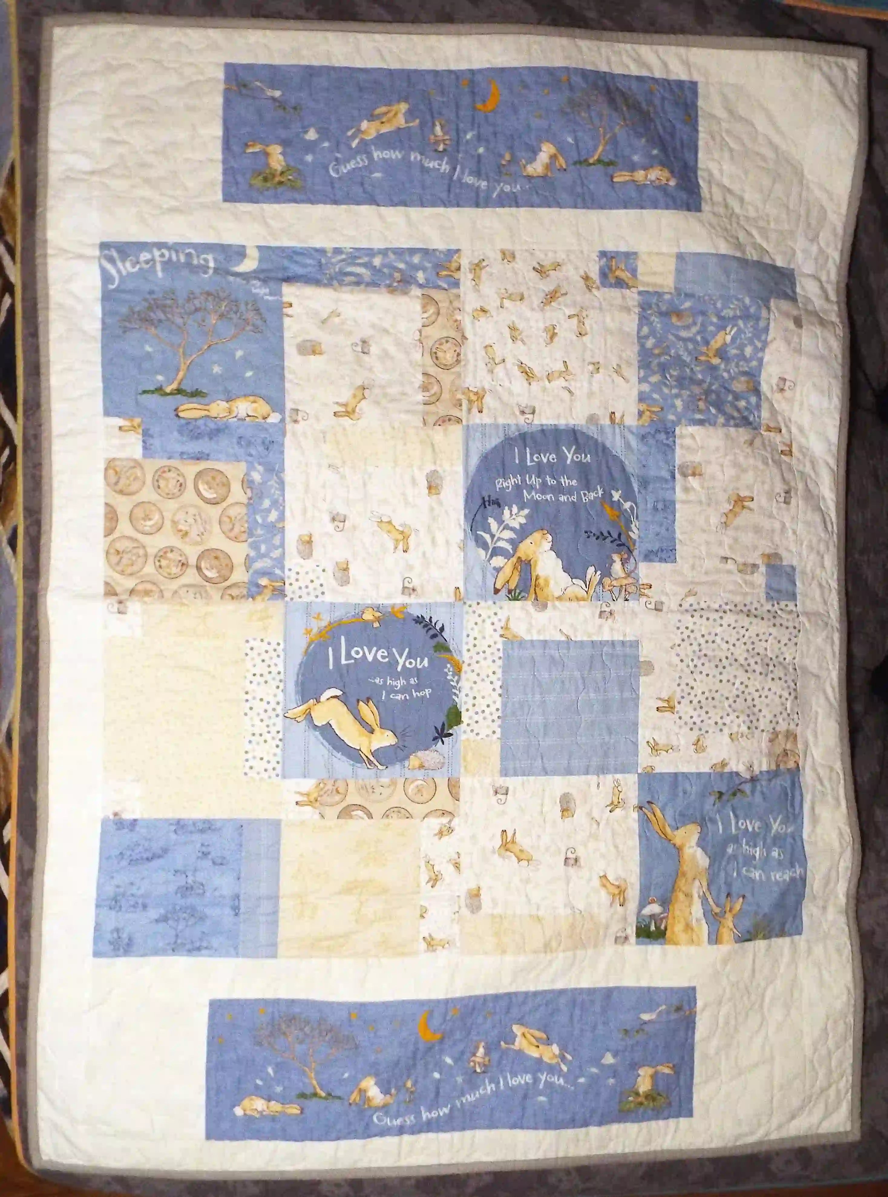 Flat lay of a handmade baby quilt with soft blue, cream, and tan bunny prints in a classic square pattern, captured in natural light on a dark gray background