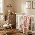 Blushing Pathways Baby Quilt shown draped over a white crib in a cream nursery.