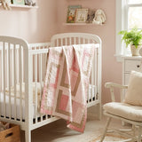 Blushing Pathways Baby Quilt shown draped over a white crib in a pale pink nursery.