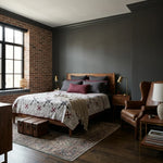 A styled bedroom interior featuring a wooden bed with Cartographer's Notes Queen Quilt, set against a dark charcoal wall and exposed brick accent wall. A brown leather wingback chair, wooden nightstands, lamps, and a patterned area rug complete the room.