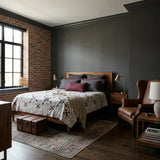 A styled bedroom interior featuring a wooden bed with Cartographer's Notes Queen Quilt, set against a dark charcoal wall and exposed brick accent wall. A brown leather wingback chair, wooden nightstands, lamps, and a patterned area rug complete the room.