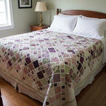 Lavender and green handmade quilt on a wooden bed with warm farmhouse decor, brass lamps, and gentle window light.