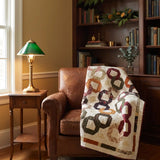 Fireside Links Lap Quilt shown on a brown leather chair in a reading nook with wooden bookshelves behind it.
