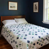 Handmade blue and green quilt shown in a dark minimally decorated room.