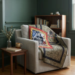 A living room scene with a light beige armchair draped with Marine Eagle Handmade Throw Quilt. A small wooden side table with a vase and book sits beside the chair, and a wooden bookcase is in the background against dark green walls.