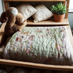 Folded green baby quilt with storybook bunny print, styled on a nursery bench with plush toys