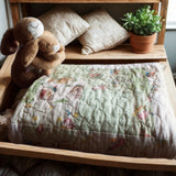 Folded green baby quilt with storybook bunny print, styled on a nursery bench with plush toys