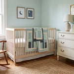 A light wood crib with Moonlit Pathways Baby Quilt draped over the side, set in a nursery with a white dresser, rocking chair, and light blue walls.