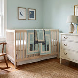 A light wood crib with Moonlit Pathways Baby Quilt draped over the side, set in a nursery with a white dresser, rocking chair, and light blue walls.
