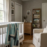A styled nursery interior featuring a white wooden crib with Moonlit Pathways Baby  Quilt draped over the side, a beige armchair, a wooden dresser, and a tall bookshelf in a gray-walled room.