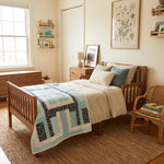 A wooden child's bed in a cozy, sunlit bedroom, styled with Moonlit Pathways Baby Quilt, beige and blue pillows, a woven jute rug, and a wicker chair with books.