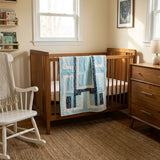 A wooden baby crib in a warm wood finish, styled in a cozy nursery with Moonlit Pathways Baby Quilt draped over the side, a white rocking chair, a wooden dresser, and a textured jute rug.