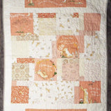 Flat-lay view of a handmade baby quilt featuring soft peach, cream, and gray blocks with gentle hare illustrations, showcasing the full patchwork design and overall texture.