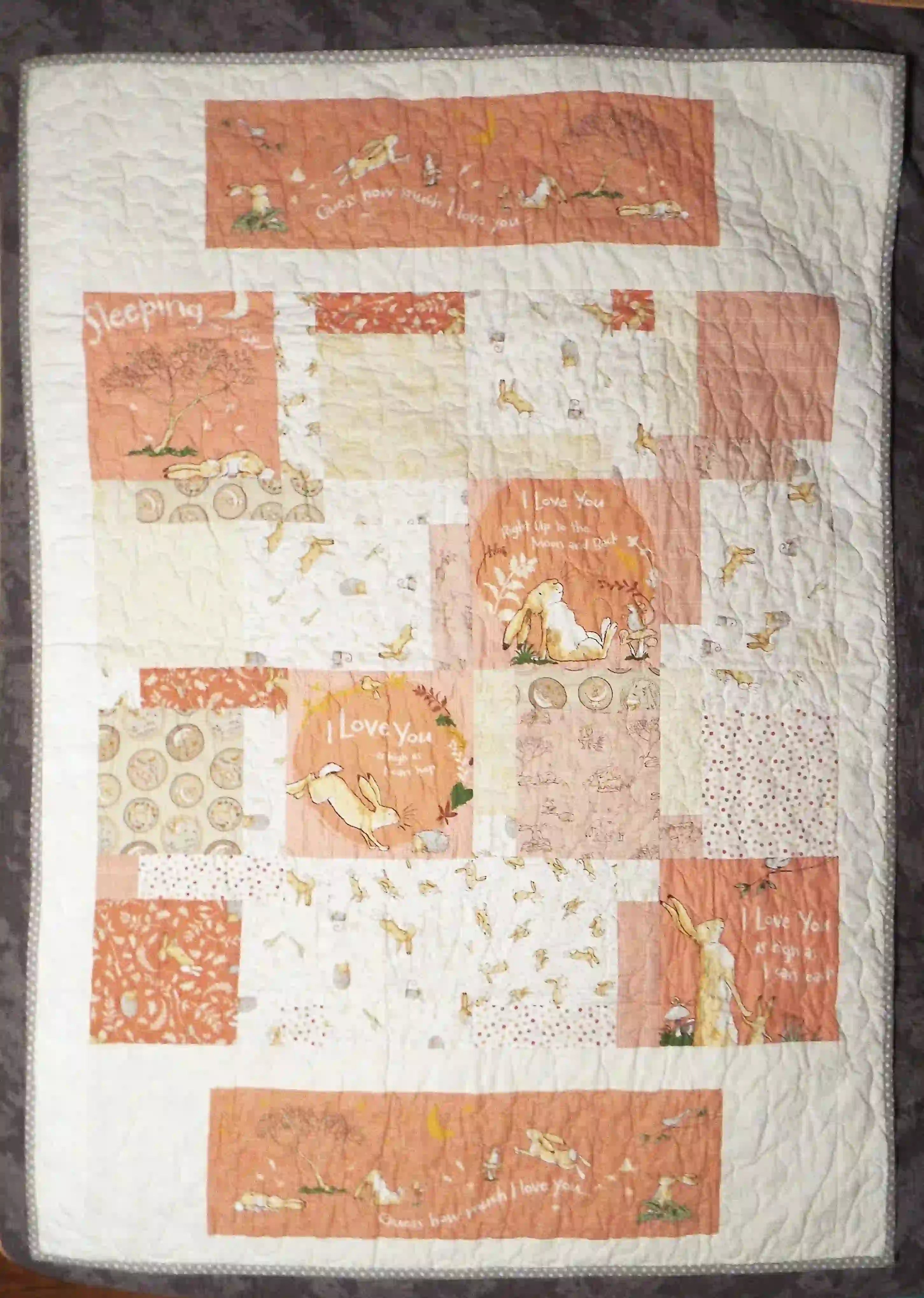 Flat-lay view of a handmade baby quilt featuring soft peach, cream, and gray blocks with gentle hare illustrations, showcasing the full patchwork design and overall texture.