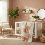 Handmade quilt draped over a modern white crib in a sunlit nursery with soft peach walls, plants, and a round wood-framed mirror, showing how the quilt pairs with warm, natural décor.