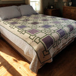 Handmade purple throw quilt spread across a queen-size bed in a cozy sunlit bedroom with wooden furniture and soft lighting.