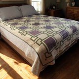 Handmade purple throw quilt spread across a queen-size bed in a cozy sunlit bedroom with wooden furniture and soft lighting.