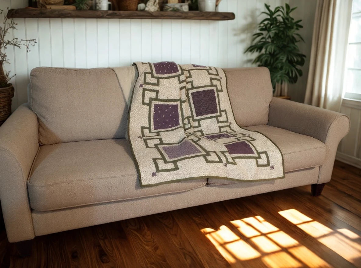 Handmade purple and cream throw quilt draped over a neutral sofa with modern farmhouse décor and a potted plant in the background.