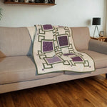 Purple and cream handmade quilt casually displayed on a beige sofa in a bright room with wood floors, white walls, and decorative shelf.