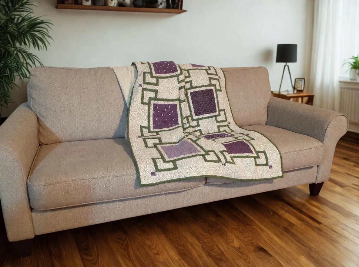 Purple and cream handmade quilt casually displayed on a beige sofa in a bright room with wood floors, white walls, and decorative shelf.