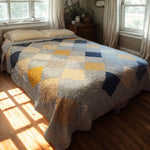 Handmade queen quilt in sunlight with blue, cream, and gold patchwork squares, perfect for cozy layered bedding.