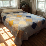 Handmade queen quilt in sunlight with blue, cream, and gold patchwork squares, perfect for cozy layered bedding.