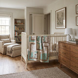 A styled nursery interior featuring a white wicker crib with Stories in the Woods Baby Quilt draped over the side, a wooden dresser, a cream armchair, and a bookshelf in a bright, neutral-toned room.