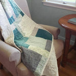 Handmade teal, cream, and gray throw shown over an upholstered chair in a reading corner
