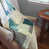 Handmade teal, cream, and gray throw shown over an upholstered chair in a reading corner