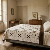 A rustic bedroom scene featuring a bed dressed in The Archivist Queen Quilt, styled with matching cream pillows and a wooden headboard.