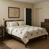 A styled bedroom featuring a bed with The Archivist Queen Quilt. The room has beige walls, a dark wood nightstand and dresser, a woven jute rug, and a framed map on the wall.