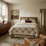 A styled bedroom interior featuring a bed dressed in The Archivist Queen Quilt, surrounded by neutral pillows. The room has warm wood floors, a matching wood dresser, a large window with sheer curtains, and a brown leather armchair in the foreground.