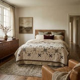 A styled bedroom interior featuring a bed dressed in The Archivist Queen Quilt, surrounded by neutral pillows. The room has warm wood floors, a matching wood dresser, a large window with sheer curtains, and a brown leather armchair in the foreground.