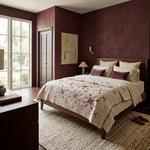 A styled bedroom interior with a bed dressed in Scarlet Correspondence Queen Quilt, set against textured dark burgundy walls. The room includes a dark wood nightstand with a lamp, a large window, and a textured beige rug.