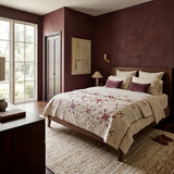 A styled bedroom interior with a bed dressed in Scarlet Correspondence Queen Quilt, set against textured dark burgundy walls. The room includes a dark wood nightstand with a lamp, a large window, and a textured beige rug.